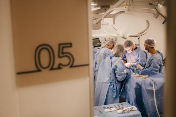 On the left is a number 5 indicating an operating theatre number, and the right has four durgeons each wearing blue scrubs from a distance in a surgical environment 