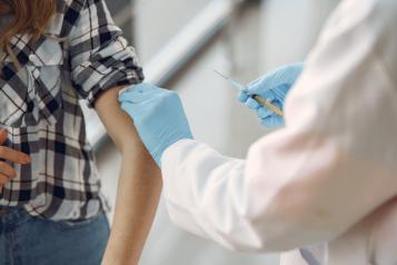 A woman in a black and white checked shirt with her sleeve rolled up and a doctor holding a vaccine syringe wearing blue surgical gloves