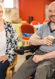 A woman sits next to a man in a wheelchair. He is holding a cup and smiling at the camera.