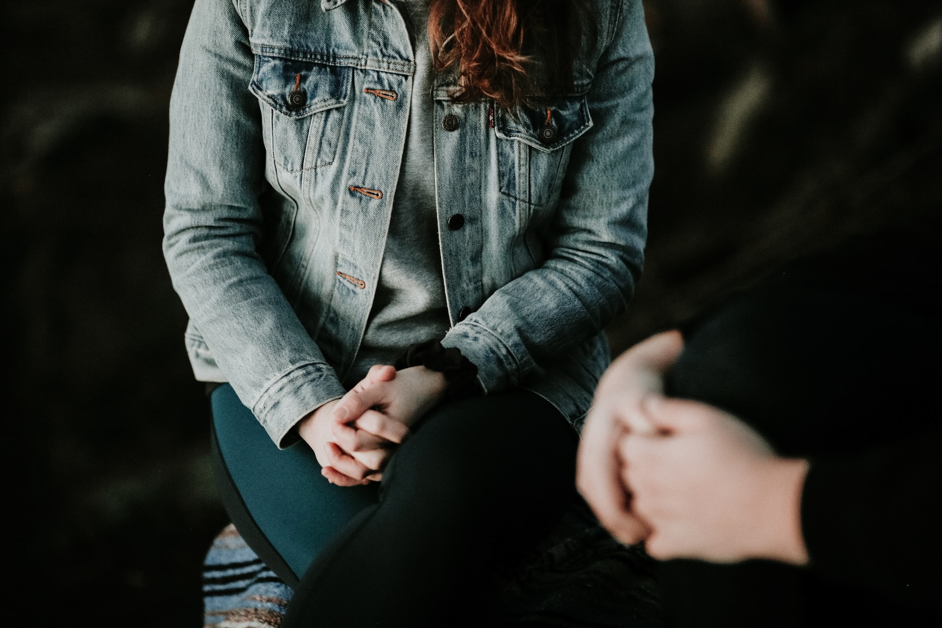 A women wearing a denim jacket sitting down, clasping her hands together. You can see the hands of the person she is talking to in the bottom right hand corner. The lighting is dark and suggests the photo is in a quiet, safe space.