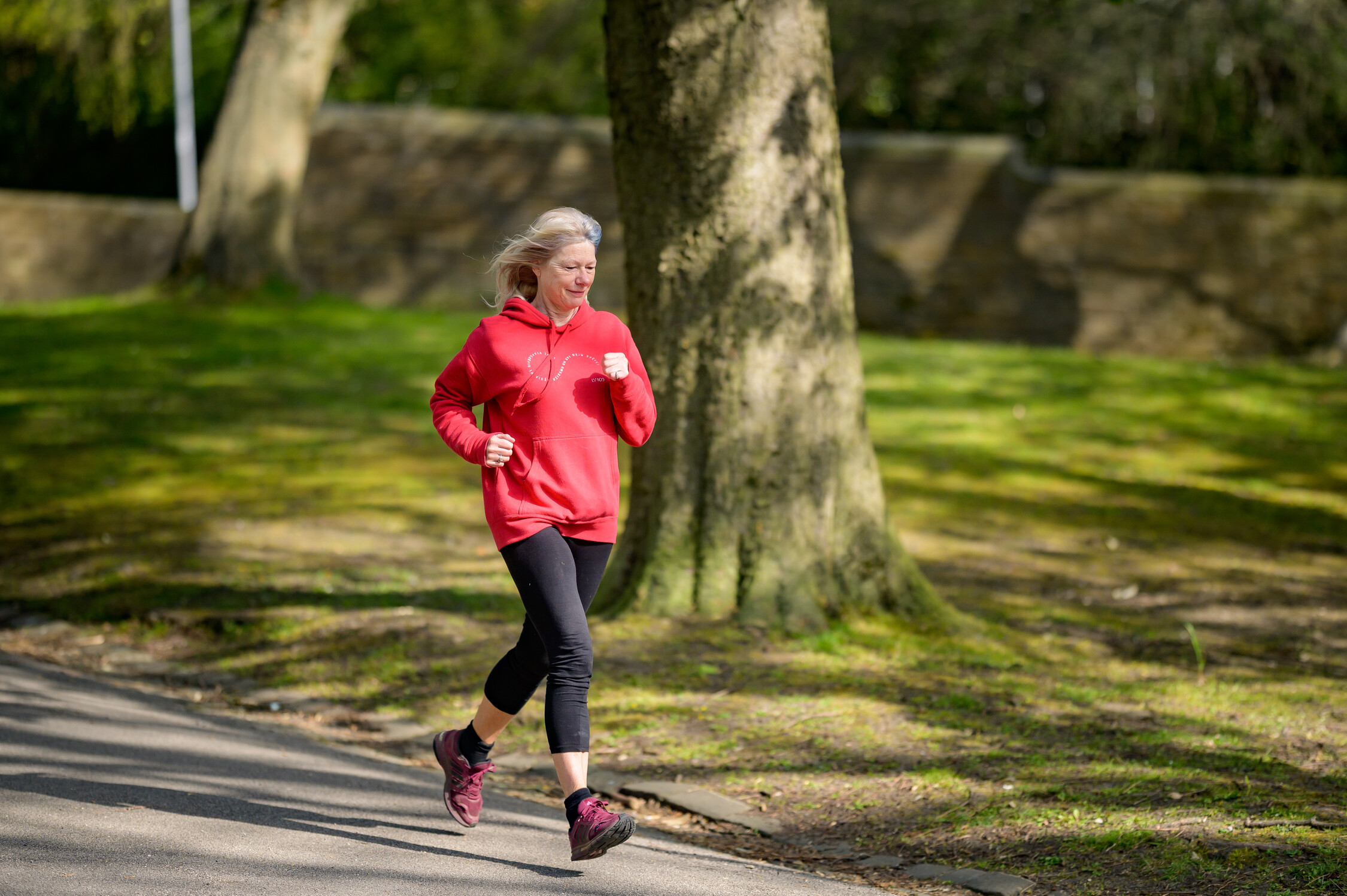 a woman with grey hair running through a park. she is wearing a red hoodie and black leggings.