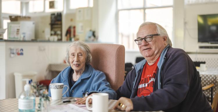An elderly man and woman sit next to each other, at a table, with mugs in front of them and a kitchen unit behind them.