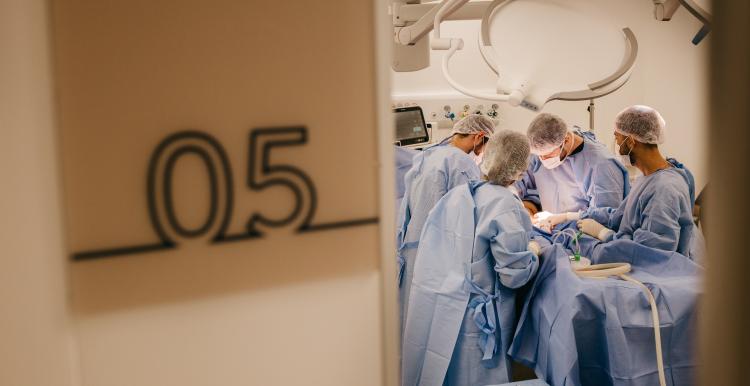 On the left is a number 5 indicating an operating theatre number, and the right has four durgeons each wearing blue scrubs from a distance in a surgical environment