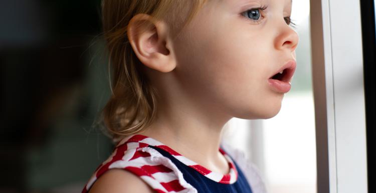 A blonde haired child in a blue, red and white dress staring out of a window