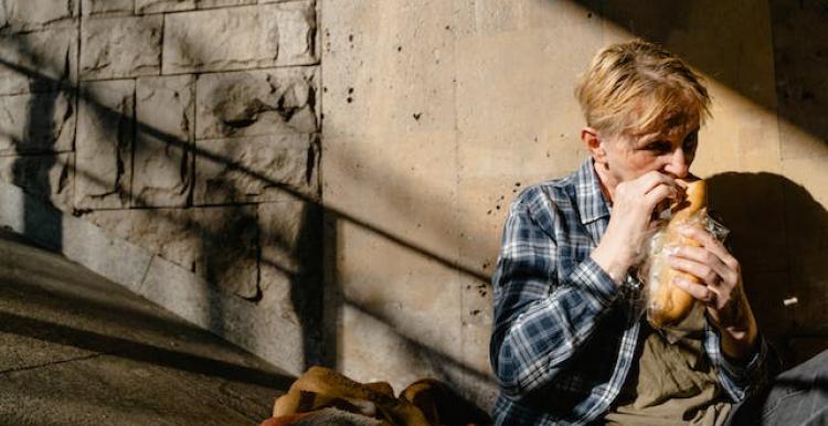 A Person in Plaid Long Sleeves Sitting on the Street while Eating Bread