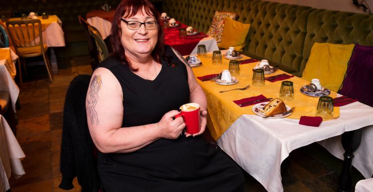 A trans woman sitting down at a table that has a yellow tablecloth. She is holding a red mug and smiling at the camera