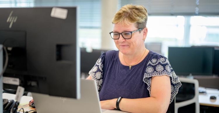 A woman with short blonde hair and glasses is sitting in front of a computer, she is slightly smiling. 