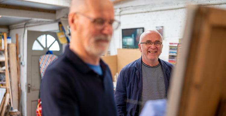 Two men standing in a room with a front door behind them. The one closest is out of focus, the man furthest from the camera is smiling and in focus