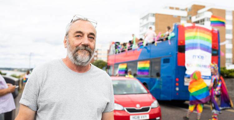 A man wearing a grey t shirt standing in front of a bus with rainbow flags 