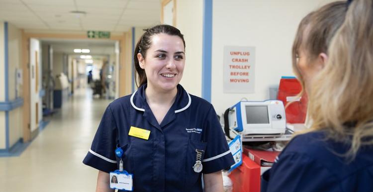 A female nurse with brown hair, wearing a navy uniform is talking to a woman with blonde hair - we can only see the back of her head. They are in a hospital corridor.