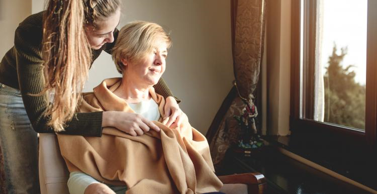 A woman is wrapped in a blanket and sits by a window with her carer