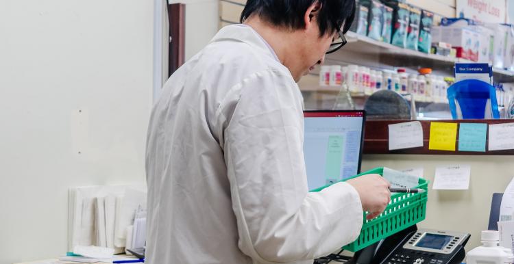A male pharmacist stands in front of a desk holding a green meds basket 