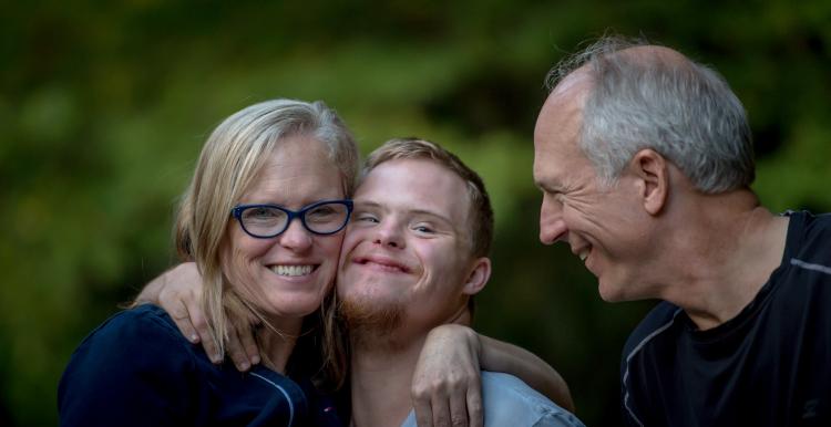 A young boy smiles at the camera and stands in between a woman and man, The woman is smiling at the camera with her arm around the boy. 