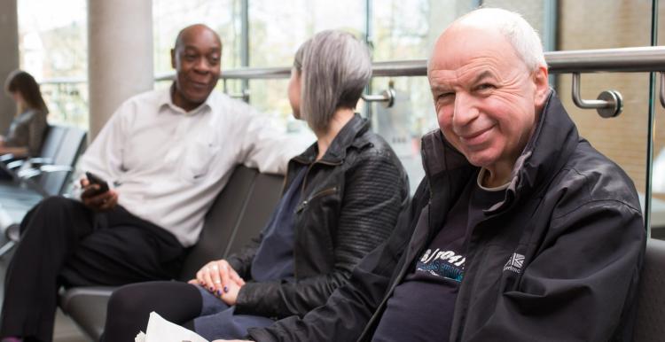 Three people sit in a waiting room. One man is holding a newspaper and smiling. Three people sit in a waiting room. One man is holding a newspaper and smiling.