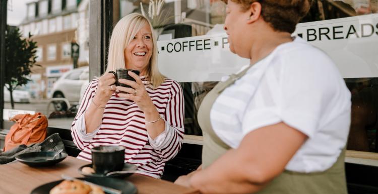 Two middle-aged women sit outside in a cafe drinking coffee, one is smiling towards the camera