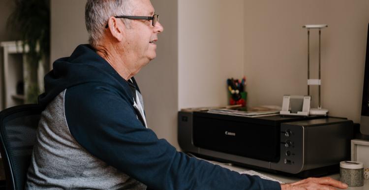 An older man wearing a blue and grey top is sitting in front of a screen as a home desk.