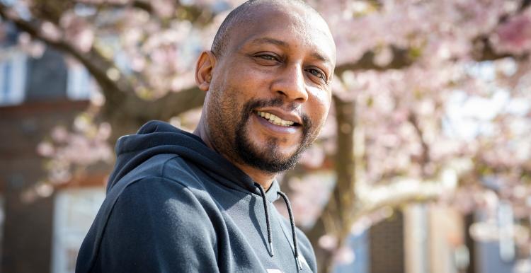 A man smiles at the camera whilst sitting outside. A blossom tree is in the background