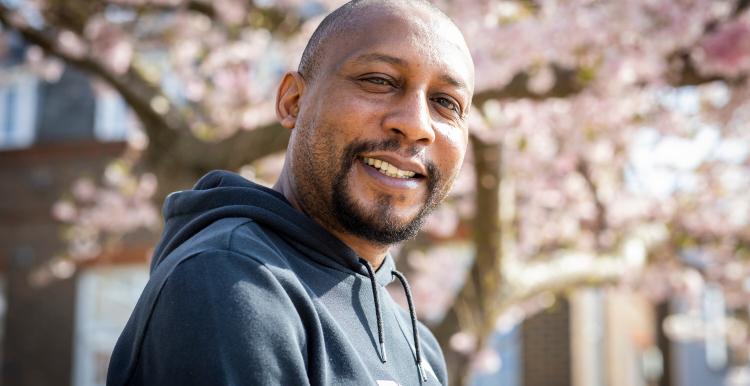 A man wearing a black Puma hoodie sitting with a pink magnolia tree behind him. He is smiling at the camera.