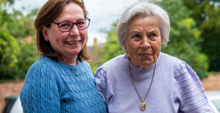 A woman with brown hair is holding the hand of an elderly lady. The woman on the left is wearing a blue jumper and smiling, the elderly lady is wearing purple and is also smiling. at the camera.