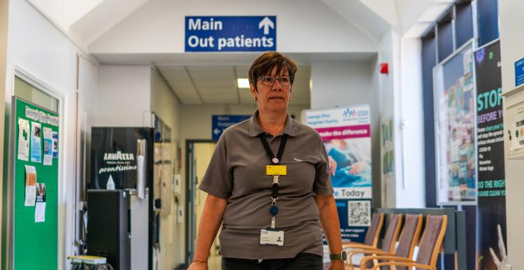 A woman with short brown hair walking along a hospital corridor. A blue sign above her head says 'Main Outpatients'