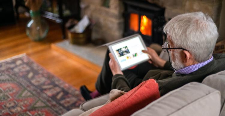 An older man wearing glasses sitting in front of a fire in a cosy-looking room, holding a laptop