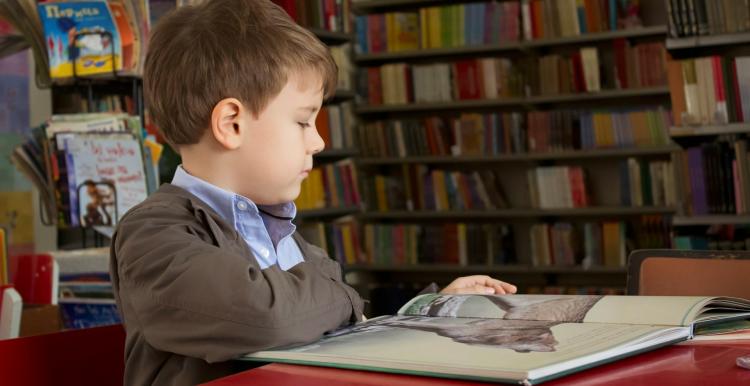 A boy surrounded by book shelves sitting down looking at a book