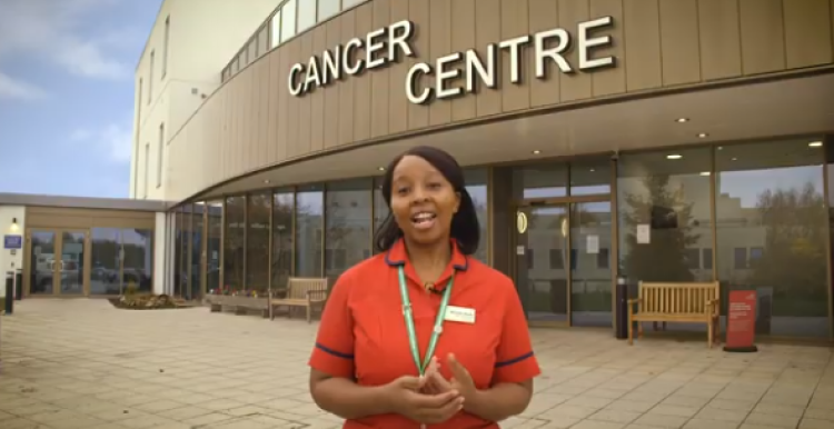 A woman wearing an orange top standing outside a building with a sign above her that says 'Cancer Centre' 