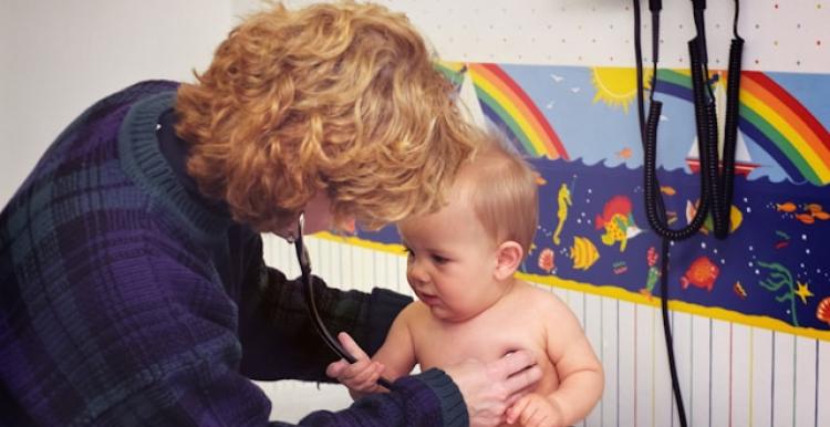 A woman is holding a baby who is sitting up in a nappy, the women is wearing a stethoscope. A rainbow print is in the background.