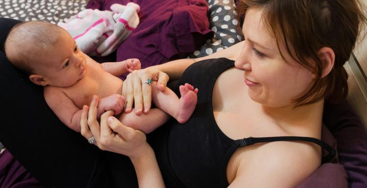 A woman with brown hair wearing a black vest lying down with a newborn baby in her lap. She is surrounded by spotty and pink blankets. She has a slight smile on her face and looks content.