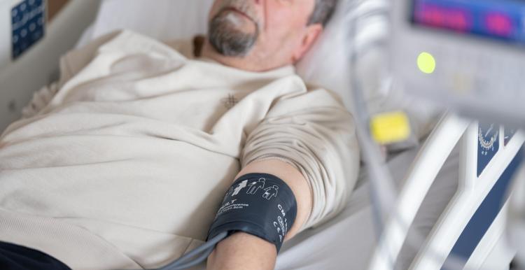 A man with a grey beard, wearing a beige t shirt lying in a hospital bed with his eyes closed. He has a blood pressure cuff on his arm.