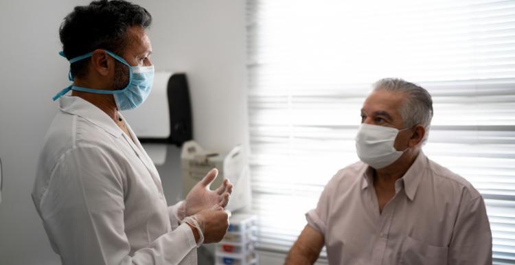 Male patient sitting on a bed talking to a male doctor. Both are wearing masks.
