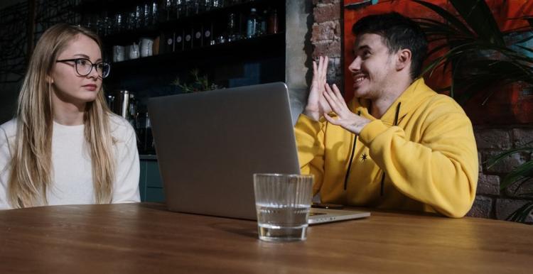 A woman on the left and a may on the right who is signing. He is wearing a yellow hoodie, sitting in front of a laptop.