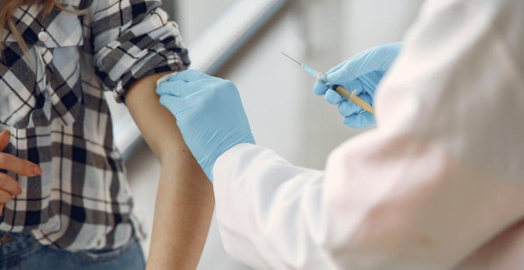 A woman in a black and white checked shirt with her sleeve rolled up and a doctor holding a vaccine syringe wearing blue surgical gloves