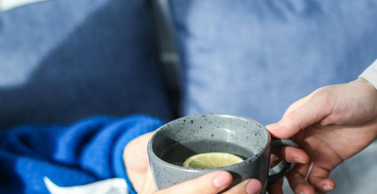 Close up of a person wearing a blue jumper holding a tissue, and someone giving them a mug with hot water and lemon. 