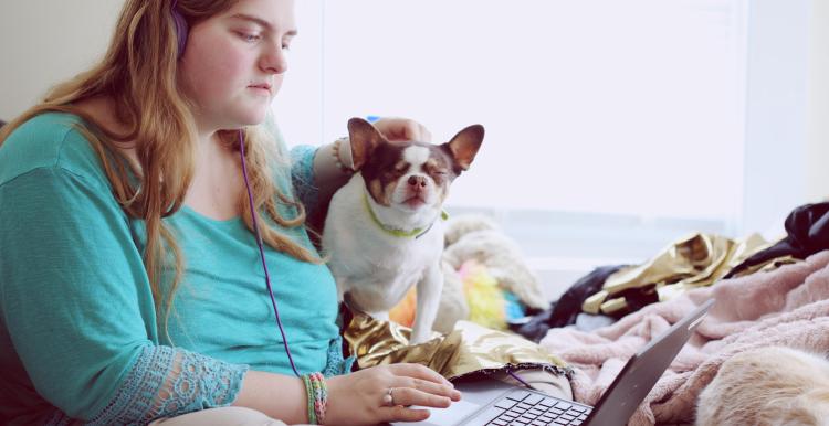 A woman wearing a turquoise top sitting down with her laptop. She is wearing headphones. A white and brown dog is sitting next to her. 