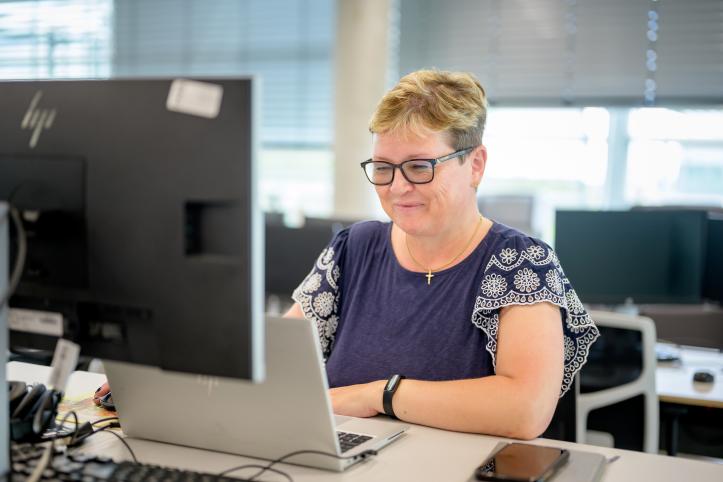 A woman with short blonde hair and glasses is sitting in front of a computer, she is slightly smiling. 
