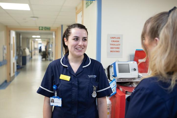A female nurse with brown hair, wearing a navy uniform is talking to a woman with blonde hair - we can only see the back of her head. They are in a hospital corridor. 