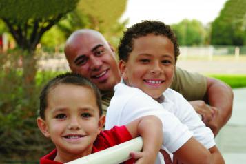 Two boys lean over a fence outside with their Dad