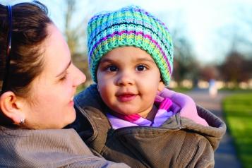 A baby in a knitted hat is held by her mum and looks at camera 