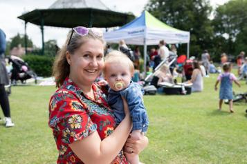 A woman stands outside holding a small baby. She is wearing a floral red dress. There is a pagoda behind her and other young children playing.