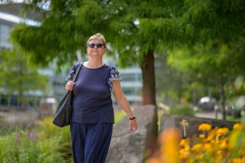 A woman wearing grey short-sleeved top and trousers is walking outside. Yellow flowers are on the right hand side and a green tree is in the background. The woman is wearing sunglasses.