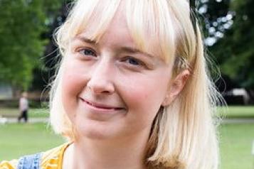 A woman with blonde hair wearing a stripey yellow top, with sunglasses on her head. She is smiling and standing in front of a green lawn and trees.