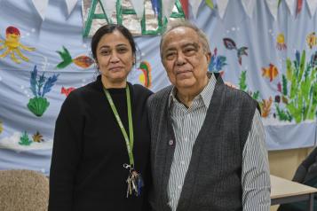 An Asian woman and older man stand side by side. She is wearing a black jumper with a lanyard around her neck, he is wearing a grey shirt and knitted waistcoat. The background behind them is bright blue with hand painted flowers.