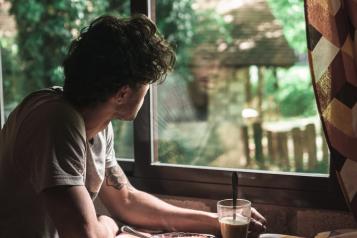 A young person looking out of a window, wearing a t shirt, and a glass and plate in front of him on a table