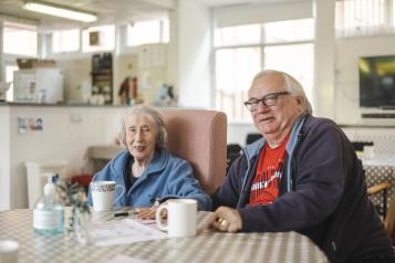 An elderly man and woman sit next to each other, at a table, with mugs in front of them and a kitchen unit behind them.