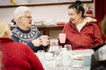 Two older women sit at a table. Both are holding a mug, they are talking. One has white hair and a blue jumper, the other had brown hair and a red coat. 