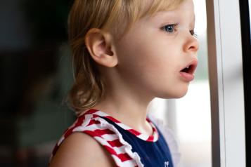 A blonde haired child in a blue, red and white dress staring out of a window