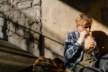 A Person in Plaid Long Sleeves Sitting on the Street while Eating Bread