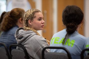 Three young girls with their backs to the camera, on the left a girl with blonde hair in plaits and a grey hoodie is talking to another girl on her right hand side, who is wearing a light blue t shirt.