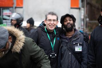 An older white man and younger black stand side by side outside smiling at the camera. They are wearing coats and name lanyards. It looks cold.  